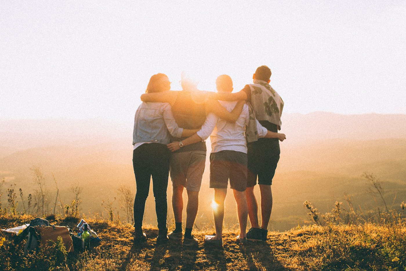 Four friends with arms around each other, watching the sun set over the hills