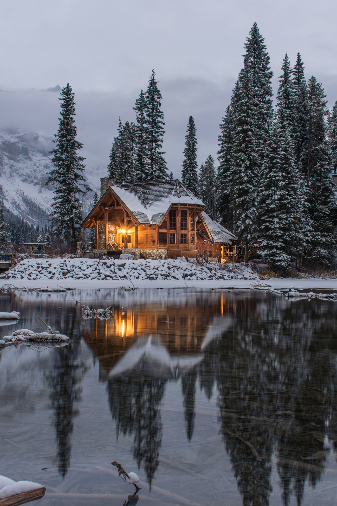 A lodge with warm, lit windows among snowy pines at dusk, reflected in a still lake